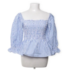 Blårandig blus med smock och volang - Säljer en söt blårandig blus med smockad överdel och volang nertill. Blusen har långa ärmar med resår och volang vid ärmslut samt fyrkantig ringning. Perfekt för vår och sommar!