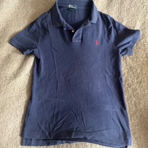 Mörkblå pikétröja från Ralph Lauren - Klassisk mörkblå pikétröja från Ralph Lauren med röd broderad logga på bröstet. Tröjan har korta ärmar, krage och knäppning med två knappar. Perfekt för en stilren och avslappnad look.