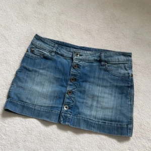 Blå jeanskjol med knappar framtill - Säljer en klassisk blå jeanskjol från only med knappar framtill och femficksmodell. Kjolen har en kort passform och är perfekt för en avslappnad stil. Snygga slitningar och detaljer på fickorna bak. Köpt här på Plick och lappen är borta men skulle uppskatta storlek 36-38