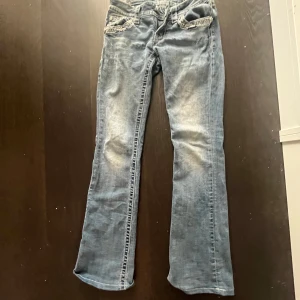 Blå bootcut jeans från Miss Me med detaljer - Säljer ett par blå bootcut jeans från Miss Me med slitningar och dekorativa sömmar. Jeansen har snygga detaljer med nitar och broderier på fickorna samt en metallpatch bak. Perfekta för dig som gillar statement-jeans!