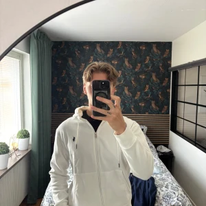Polo Ralph Lauren Zip Tröja  - Säljer en stilren vit hoodie från Polo Ralph Lauren med dragkedja och huva. Tröjan har två fickor framtill och den klassiska broderade loggan på bröstet. Perfekt för en avslappnad och snygg look.