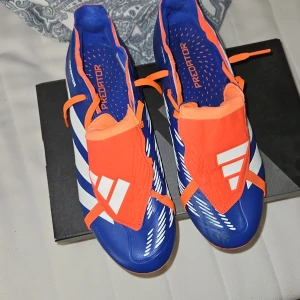 Adidas Predator fotbollsskor blå och orange - Säljer ett par Adidas Predator fotbollsskor i blått och orange med vita detaljer. Skorna har snörning och är designade för spel på gräs med fasta dobbar. Perfekta för dig som vill sticka ut på planen!