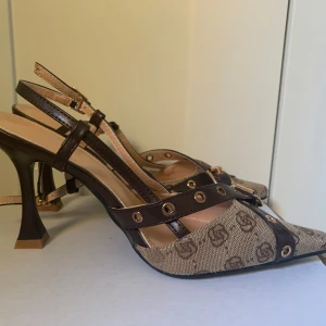 Bruna pumps med mönstrad canvas, remmar och guldetaljer - Säljer ett par snygga pumps med spetsig tå och klack. Skorna har bruna läderremmar med guldfärgade detaljer och canvas med mönster. Perfekta för dig som vill sticka ut med en unik design och cool form på klacken. Har aldrig används. 