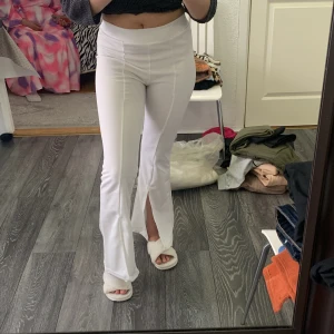 Vita utsvängda byxor med slits - Säljer ett par vita utsvängda byxor med hög midja och slits framtill vid bensluten. Byxorna har en tight passform upptill och är tillverkade i ett stretchigt material som sitter snyggt på kroppen.