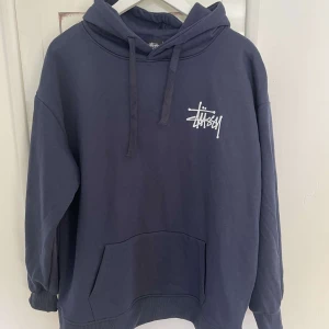 Mörkblå hoodie från Stüssy - Säljer en mörkblå oversized hoodie från Stüssy med vit logga på bröstet och stor logga på ryggen. Perfekt för en avslappnad streetwear-look.