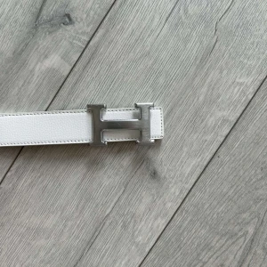Vitt bälte med H-spänne - Snyggt vitt bälte med ett stort H-format spänne i silverfärg. Bältet har en stilren design och är tillverkat i skinn med vita sömmar längs kanterna. Perfekt accessoar för att lyfta din outfit.