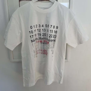 Vit t-shirt från Maison Margiela - Vit t-shirt med svart tryck framtill från Maison Margiela. Trycket består av siffror och text, med en del av trycket i rött och en cool, droppande effekt. 