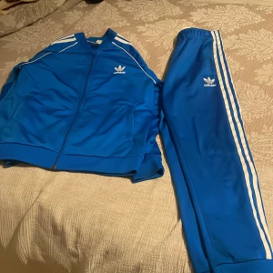 Blå Adidas träningsbyxor med vita ränder - Säljer ett par klassiska blå Adidas träningsbyxor med vita ränder längs sidorna och Adidas-logga på låret. Byxorna har elastisk midja och är perfekta för sport eller chill. Matchar med tillhörande jacka om du vill köra hela looken.