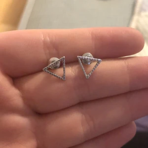 Triangelformade örhängen i silverfärg - Säljer ett par stilrena örhängen i form av öppna trianglar med små glittrande stenar. De är i silverfärg och har klassisk plupp-bakdel. Perfekta för dig som gillar minimalistisk design.