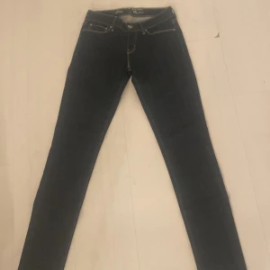 Mörkblå Levi's Demi Curve jeans - Snygga mörkblå Levi's Demi Curve jeans med klassisk femficksdesign och smala ben. Byxorna har guldfärgade sömmar och knappstängning framtill. Perfekta för dig som gillar en tidlös och stilren look.