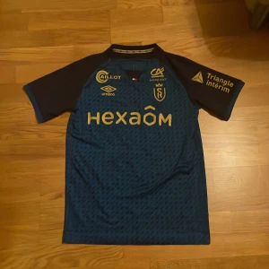 Blå fotbollströja Stade de Reims Flips 12 från Umbro - Säljer en blå fotbollströja från Stade de Reims med guldiga detaljer och tryck, spelarnamn Flips och nummer 12 på ryggen. Tröjan har korta ärmar och är tillverkad i ett lätt och ventilerande material. Perfekt för fotbollsfans!