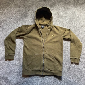 Olivgrön hoodie från Lyle & Scott - Säljer en olivgrön hoodie från Lyle & Scott Junior med dragkedja och huva. Tröjan har två fickor framtill och en broderad logga på bröstet. Perfekt för en avslappnad stil. Nypris 699kr mitt pris 199kr