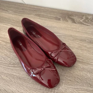 Vinröda ballerinaskor  - Säljer ett par glansiga vinröda ballerinaskor med söt rosettdetalj framtill, perfekta för en stilren och klassisk look.