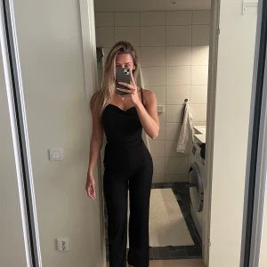 Svart byxdress med smala axelband - Säljer en svart byxdress med smala axelband och snygg omlottdetalj framtill. Byxdressen har öppen rygg med knyt och långa, raka ben. Perfekt för dig som vill ha en stilren och trendig look.