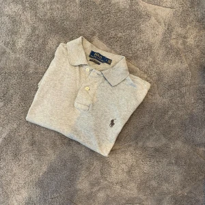 Grå pikétröja från Polo Ralph Lauren - Säljer en klassisk grå pikétröja från Polo Ralph Lauren i bomull. Tröjan har korta ärmar, krage och knäppning med två knappar framtill. Ikoniska broderade loggan på bröstet. Perfekt till vardags och enkel att matcha.