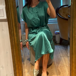 Grön klänning i satin - Säljer en elegant grön klänning i satin med knyt i midjan och korta ärmar. Klänningen har en avslappnad passform och faller mjukt ner till vaden. Perfekt för dig som gillar stilrena och bekväma plagg. Ena öglan trasig vid köp men har inte påverkats av det