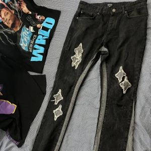 Svarta jeans med slitningar från BoohooMAN - Säljer ett par svarta jeans från BoohooMAN med unika slitningar och trasiga detaljer på benen. Byxorna har raka ben och en klassisk femficksdesign. Perfekta för dig som gillar en edgy streetstil.