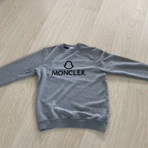 Grå sweatshirt från Moncler - Säljer en stilren grå sweatshirt från Moncler med svart logotyp och tryck på bröstet. Tröjan har rund halsringning, långa ärmar och ribbade muddar vid ärmslut och nederkant. Perfekt för en avslappnad och trendig look. 