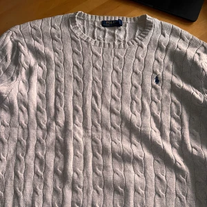 Beige kabelstickad tröja från Polo Ralph Lauren - Säljer en klassisk beige kabelstickad tröja från Polo Ralph Lauren. Tröjan har rund halsringning, lång ärm och den ikoniska broderade loggan på bröstet. Perfekt för en stilren och tidlös look.