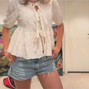 Vit blus med knyt fram - Säljer en söt vit blus med tre knyt framtill och lätt volang nertill. Blusen har korta ärmar och ett subtilt mönster i tyget som ger en fin struktur. Perfekt till både jeans och kjol.