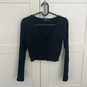 Svart ribbad croppad omlottopp - Säljer en svart croppad topp med omlott-skärning och djup v-ringning. Toppen är ribbad och har långa ärmar, perfekt för en trendig och stilren look. Toppen är från New Yorker och är i storlek S.