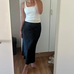 Svart långkjol i satin - Säljer en svart långkjol i satin med lätt glansig finish. Kjolen har en rak och smal passform som faller snyggt längs kroppen. Perfekt att styla med en enkel topp eller stickad tröja. Elastisk midja