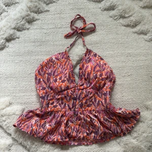 Blommig halterneck topp från TEN11 - Säljer en färgglad halterneck topp från TEN11 i storlek XS. Toppen har ett blommigt mönster i orange, rosa och lila toner, med volang nertill och smockad rygg. Perfekt för varma dagar och somriga outfits! Unik från Cambodja, använd en enstaka gång säljer pågrund av för liten storlek.