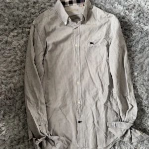 Randig skjorta från Burberry - Säljer en klassisk vit och grå smalrandig skjorta från Burberry med knappar framtill och diskret broderad logga på bröstet. Skjortan har lång ärm och är tillverkad i 100% bomull. Perfekt för dig som gillar stilrena plagg med exklusiv känsla.