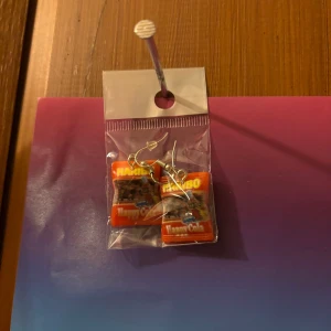 Haribo örhängen med colagodis - Unika örhängen med små Haribo cola-godis som hängsmycke. Perfekt för dig som gillar roliga och färgglada accessoarer. Örhängena har silvriga krokar och detaljerad design som ser ut som godisförpackningar.