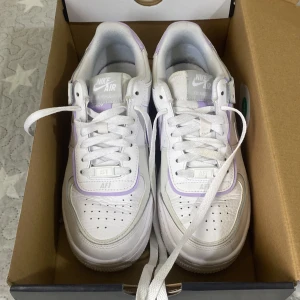 Nike Air Force 1 Shadow vit/lila - Nike Air Force 1 Shadow i vitt med lila detaljer. Klassisk chunky sula, perforerad tå och dubbla lager på swoosh och häl. Skorna har snörning och är tillverkade i syntetmaterial och skinn. Perfekta för dig som vill ha en unik twist på en ikonisk sneaker.
