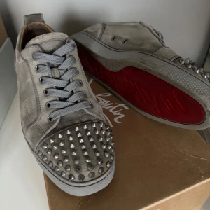 Gråa sneakers med nitar från Christian Louboutin - Säljer ett par gråa sneakers från Christian Louboutin med grå snörning och silverfärgade nitar på tån. Skorna har den klassiska röda sulan och är tillverkade i mocka med detaljer i skinn på insidan. Og all, skick 7/10 (alla nitar kvar men lite heel drag). Perfekta till våren/sommaren!