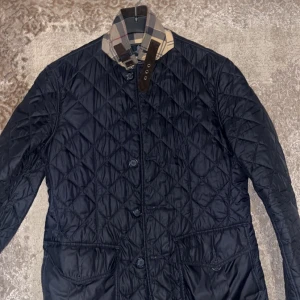 Svart barbour jacka, unisex passar storlek m/L - Säljer en svart quiltad jacka från Burberry med klassiskt rutigt foder och krage. Jackan har knappar framtill, två fickor och snyggt bälte vid halsen. Perfekt för dig som vill ha en stilren och tidlös look.