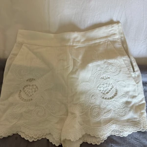 Vita spetsdetaljerade shorts från Zara - Säljer ett par vita shorts från Zara med vackra broderade spetsdetaljer och vågig kant nertill. Shortsen har fickor fram och är tillverkade i ett lätt linnematerial, perfekta för varma dagar. Aldrig använda