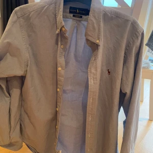 Ljusblå skjorta från Ralph Lauren - Klassisk ljusblå skjorta från Ralph Lauren med knappar framtill och broderad logga på bröstet. Skjortan har lång ärm och normal passform, perfekt till många olika tillfällen. Storleken är custom fit sitter som en s/m