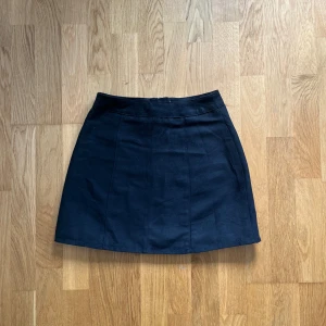 Svart kortkjol från Divided - Säljer en svart kortkjol från Divided med dragkedja bak. Kjolen har en enkel och stilren design som passar till många olika outfits. Materialet känns mjukt och har en lätt struktur.