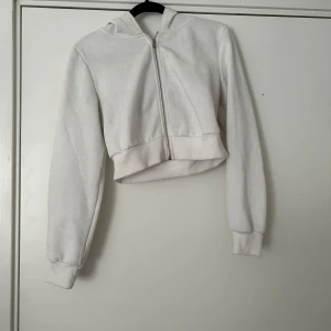 Vit croppad hoodie med dragkedja - Säljer en vit croppad hoodie med dragkedja framtill och huva. Tröjan har långa ärmar och breda muddar vid ärmslut och nederkant. Perfekt för en avslappnad stil.