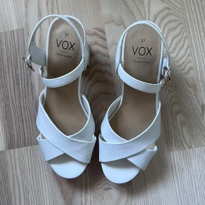Vita platåsandaler från VOX - Säljer ett par vita sandaler från VOX med hög klack och platåsula. Skorna har öppen tå, breda korsade remmar framtill och justerbart spänne vid ankeln. Tillverkade i syntetmaterial och passar perfekt till sommaren. Använda 2 gånger💕