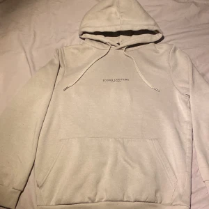 Beige hoodie från Icono Couture - Säljer en beige hoodie från Icono Couture med diskret logga framtill och större tryck på ryggen. Tröjan har huva med dragsko och en stor magficka. Perfekt för en avslappnad och stilren look.