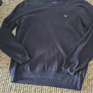 Mörkblå stickad tröja från GANT - Säljer en klassisk mörkblå stickad tröja från GANT med rund halsringning och diskret broderad logga på bröstet. Perfekt för lager-på-lager och passar till många olika stilar. Tröjan har långa ärmar och ribbade muddar.
