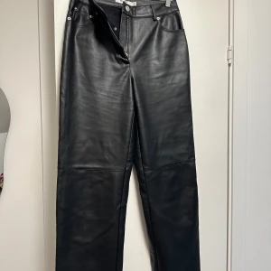 Svarta skinnbyxor från NA-KD (faux) - Snygga svarta skinnbyxor från NA-KD med raka ben och klassisk femficksdesign. Byxorna har hög midja och silverfärgade detaljer vid fickorna. Perfekta för en trendig och stilren look.