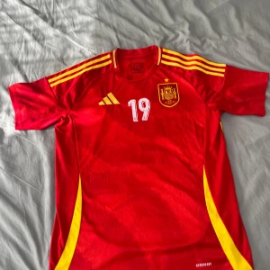 Spansk landslagströja Lamine Yamal 19 - Röd fotbollströja från Adidas med gula detaljer, nummer 19 och Lamine Yamal på ryggen. Spanska landslagets emblem på bröstet och tre gula ränder på axlarna. Perfekt för dig som vill stötta Spanien!
