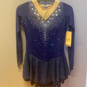 Marinblå konståkningsklänning med strass - Superfin marinblå konståkningsklänning med lång ärm och glittrande strassdetaljer framtill och baktill. Klänningen har en v-ringad mesh i beige vid halsen och en liten kjol. Perfekt för isen!