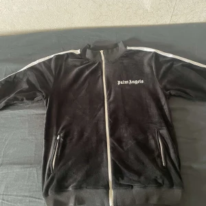 Svart track jacket Palm Angels - Säljer en svart track jacket från Palm Angels med vit dragkedja. Jackan har ståkrage, två fickor med dragkedja och logga på bröstet. Den är knappt använd och i väldigt bra skick. Den har en välvårdad velvet material.