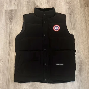 Svart dunväst från Canada Goose - Säljer en svart dunväst från Canada Goose med hög krage och klassisk logotyp på bröstet. Västen har både dragkedja och knappar framtill. västen har fem stora fixkor. Perfekt för kyliga dagar och enkel att matcha med olika outfits.