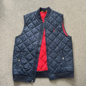 Mörkblå väst från Ralph Lauren - Säljer en mörkblå quiltad väst från Ralph Lauren med röd insida och broderad logga på bröstet. Västen har dragkedja framtill, två fickor med tryckknappar och ribbade detaljer vid krage och midja. Perfekt för en avslappnad look! Skick: 10/10. Det är Xl i barn storlek men S i vanlig storlek. Hör av dig vid andra frågor / funderingar! 