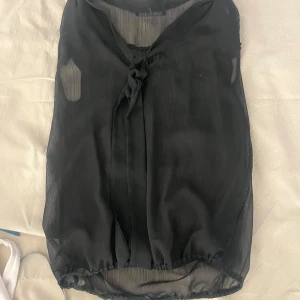 Svart blus med knyt i halsen - Säljer en svart, tunn och luftig blus med knyt i halsen. Blusen har en lätt transparent känsla och är perfekt för att skapa en stilren look. Den har normal passform och är ärmlös, vilket gör den enkel att matcha med olika outfits.