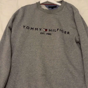 Grå sweatshirt från Tommy Hilfiger - Säljer en klassisk grå sweatshirt från Tommy Hilfiger med broderad logga och text på bröstet. Tröjan har rund halsringning och långa ärmar. Perfekt för en avslappnad och stilren look.