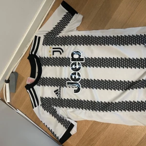 Juventus fotbollströja från Adidas - Juventus matchtröja i vitt med svarta geometriska ränder och svarta detaljer. Adidas-logga, Juventus klubbmärke och Jeep som sponsor på bröstet. Klassisk rund hals och korta ärmar. Perfekt för fotbollsfans!