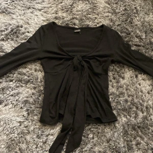 Svart knytblus från Gina Tricot - Säljer en svart långärmad topp från Gina Tricot med knyt framtill. Toppen har djup v-ringning och är i ett mjukt, stretchigt material. 