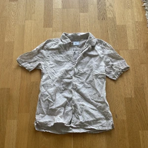 Beige linneskjorta från Bläck - Säljer en beige kortärmad skjorta från Bläck i svalt linnematerial. Skjortan har klassisk krage och knäppning framtill, perfekt för varma dagar. Enkel och stilren design som passar till mycket.